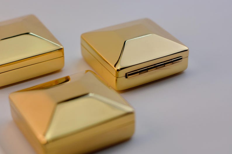 Puede incluir: Tres cajas doradas de forma cuadrada con un dise&ntilde;o geom&eacute;trico. Las cajas tienen una tapa con bisagras y una superficie brillante y reflectante. Las cajas est&aacute;n dispuestas sobre una superficie clara.