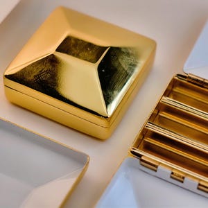 Puede incluir: Una colecci&oacute;n de cajas y bandejas geom&eacute;tricas de color dorado. La caja principal es cuadrada con una parte superior en forma de diamante. Algunas bandejas est&aacute;n abiertas, revelando un interior blanco y detalles dorados. Los art&iacute;culos est&aacute;n dispuestos sobre una superficie blanca.