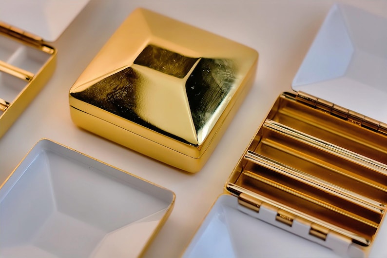 Puede incluir: Una colecci&oacute;n de cajas y bandejas geom&eacute;tricas de color dorado. La caja principal es cuadrada con una parte superior en forma de diamante. Algunas bandejas est&aacute;n abiertas, revelando un interior blanco y detalles dorados. Los art&iacute;culos est&aacute;n dispuestos sobre una superficie blanca.