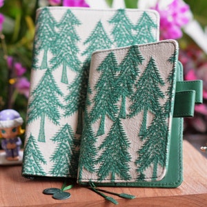 May include: Two green and white journals with a repeating embroidered pine tree pattern. One journal is larger than the other. The smaller journal has a green strap closure. The journals are on a wooden surface.