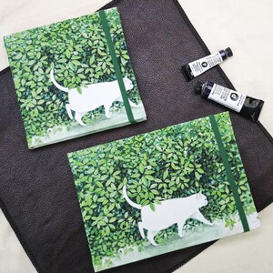 May include: Two square notebooks with a white cat design walking through green foliage. Each notebook has a green elastic band closure. Two tubes of blue paint are also visible on a dark brown surface.