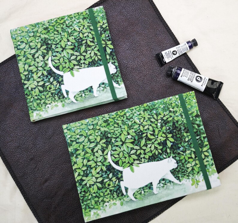 May include: Two square notebooks with a white cat design walking through green foliage. Each notebook has a green elastic band closure. Two tubes of blue paint are also visible on a dark brown surface.