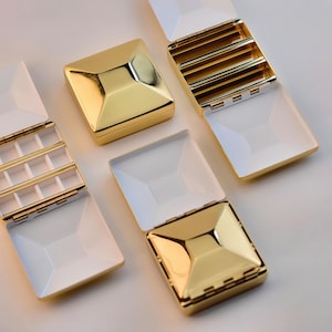 Puede incluir: Se muestran varias joyeros dorados y blancos, algunos abiertos y otros cerrados. Las cajas tienen un dise&ntilde;o geom&eacute;trico con una parte superior dorada en forma de pir&aacute;mide y una base blanca rectangular. Las cajas abiertas revelan compartimentos.