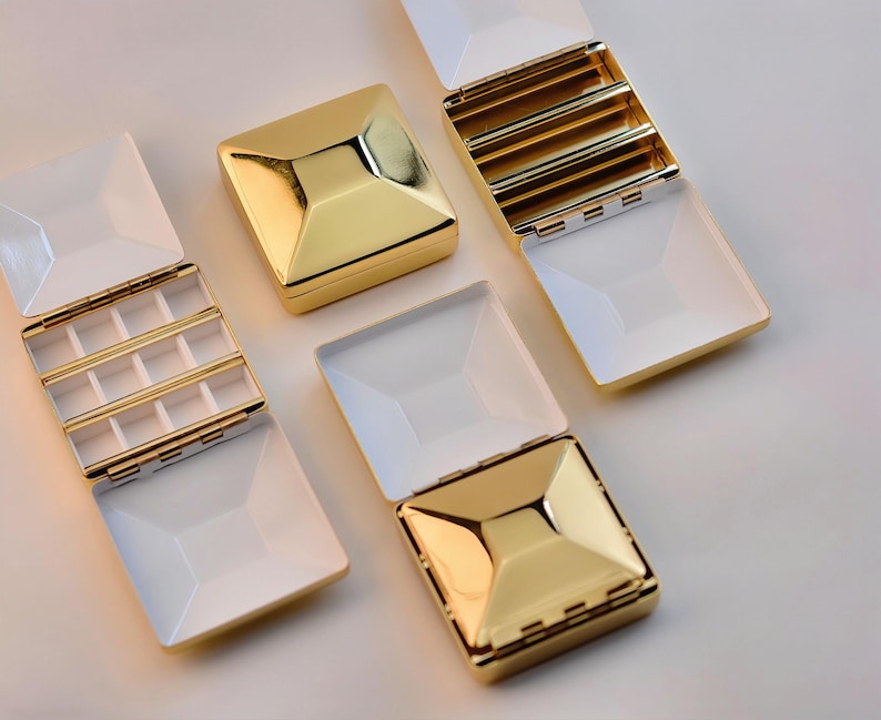 Puede incluir: Se muestran varias joyeros dorados y blancos, algunos abiertos y otros cerrados. Las cajas tienen un dise&ntilde;o geom&eacute;trico con una parte superior dorada en forma de pir&aacute;mide y una base blanca rectangular. Las cajas abiertas revelan compartimentos.