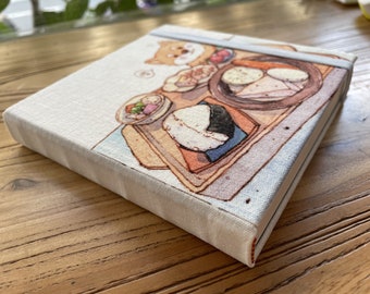 May include: A white linen notebook with a colorful illustration of a cartoon dog and a plate of Japanese food, including rice balls, vegetables, and a bowl of soup. The notebook has a red ribbon closure.