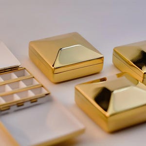 Puede incluir: Cajas de joyer&iacute;a doradas y geom&eacute;tricas. Una caja est&aacute; abierta, revelando un interior blanco con peque&ntilde;os compartimentos. Las otras cajas est&aacute;n cerradas, mostrando una superficie brillante y reflectante. Las cajas est&aacute;n dispuestas sobre un fondo claro.