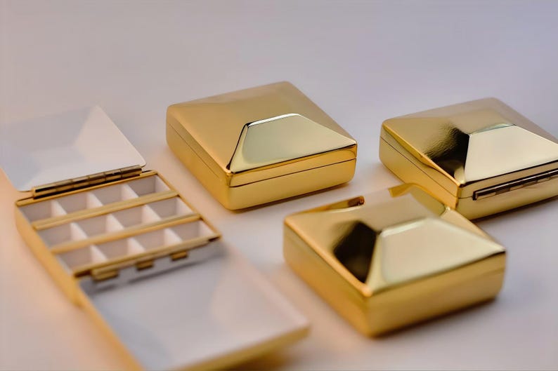 Puede incluir: Cajas de joyer&iacute;a doradas y geom&eacute;tricas. Una caja est&aacute; abierta, revelando un interior blanco con peque&ntilde;os compartimentos. Las otras cajas est&aacute;n cerradas, mostrando una superficie brillante y reflectante. Las cajas est&aacute;n dispuestas sobre un fondo claro.