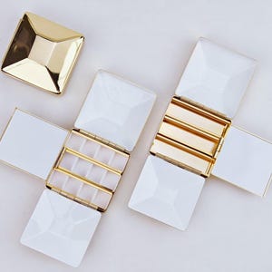 Puede incluir: Cajas de joyer&iacute;a blancas y doradas con un dise&ntilde;o geom&eacute;trico. Las cajas est&aacute;n abiertas, revelando compartimentos para guardar objetos peque&ntilde;os. Las tapas son cuadradas con forma de pir&aacute;mide. Una tapa est&aacute; separada, las otras est&aacute;n unidas.