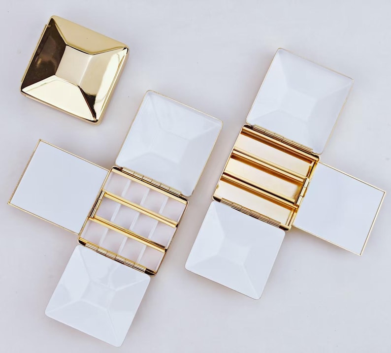 Puede incluir: Cajas de joyer&iacute;a blancas y doradas con un dise&ntilde;o geom&eacute;trico. Las cajas est&aacute;n abiertas, revelando compartimentos para guardar objetos peque&ntilde;os. Las tapas son cuadradas con forma de pir&aacute;mide. Una tapa est&aacute; separada, las otras est&aacute;n unidas.