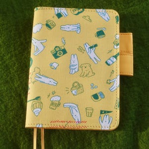 Pode incluir: Capa de caderno amarela com um padrão repetido de ilustrações de desenhos animados em verde e branco. A capa apresenta um fecho de aba e dois pingentes amarelos em forma de borla. Fundo verde texturizado.