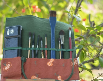 May include: A green canvas roll-up case with a brown fabric flap and a black watercolor paint set. The case has multiple slots for holding paint brushes. The case is decorated with a pumpkin pattern.