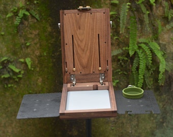 May include: Open wooden box with a light grey interior and brass fittings. The box is on a black surface with a small green bowl. The background is a wall covered in green plants.