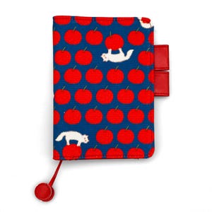 May include: A rectangular notebook cover in deep blue featuring a repeating pattern of bright red apples. Two white cats are integrated into the design, one perched on an apple and another holding one. It includes two red pen loops and a red bookmark.