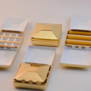 Puede incluir: Se muestran varias joyeros cuadrados de color dorado con interiores blancos. Algunas cajas est&aacute;n abiertas, revelando compartimentos. Las cajas tienen un dise&ntilde;o geom&eacute;trico y est&aacute;n dispuestas sobre una superficie blanca.