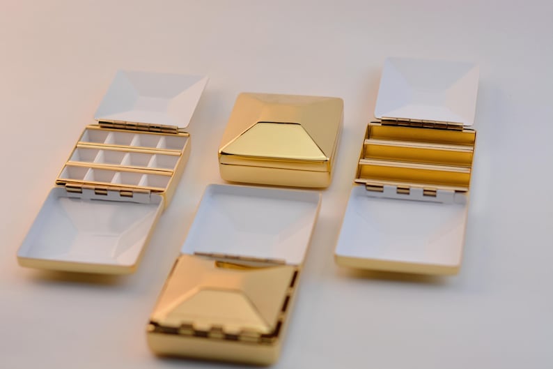 Puede incluir: Se muestran varias joyeros cuadrados de color dorado con interiores blancos. Algunas cajas est&aacute;n abiertas, revelando compartimentos. Las cajas tienen un dise&ntilde;o geom&eacute;trico y est&aacute;n dispuestas sobre una superficie blanca.