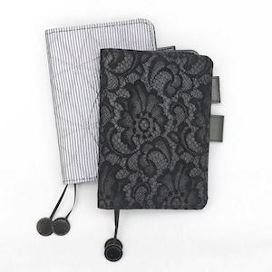 May include: Two notebooks. One has a black lace cover, the other has a white and blue striped cover. Both have black ribbon bookmarks with decorative black circles at the ends. The lace notebook has three black tabs.