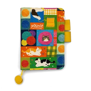 May include: A rectangular fabric notebook cover featuring a colorful patchwork design of cats and yarn balls. The cover displays the word pounce in white text, with yellow pen loops on the side and a yellow circular charm attached to a thin cord.