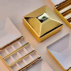 Puede incluir: Cajas de joyer&iacute;a geom&eacute;tricas doradas con interiores blancos. Algunas cajas est&aacute;n abiertas, revelando compartimentos y divisores. Las cajas tienen una superficie brillante y reflectante, y las tapas est&aacute;n articuladas. El dise&ntilde;o general es moderno y elegante.