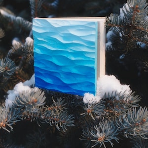 May include: A square wooden art piece with a carved wave design, painted in shades of blue. The artwork is displayed outdoors, resting on a snow-covered evergreen branch. The piece has a white border.