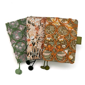 May include: Three rectangular fabric notebook covers featuring intricate floral and bird patterns. The left cover is green, the center is peach, and the right is brown. Each cover is embellished with raised beaded lattice detailing and includes matching ribbon bookmarks.