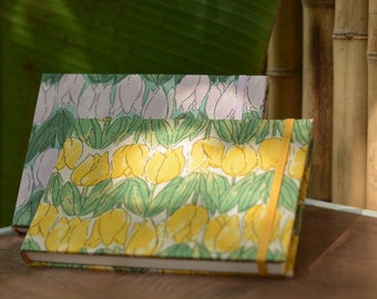 May include: Two notebooks with floral patterns. The top notebook has a light purple floral pattern. The bottom notebook has a yellow floral pattern with green leaves. Both notebooks have a yellow ribbon.