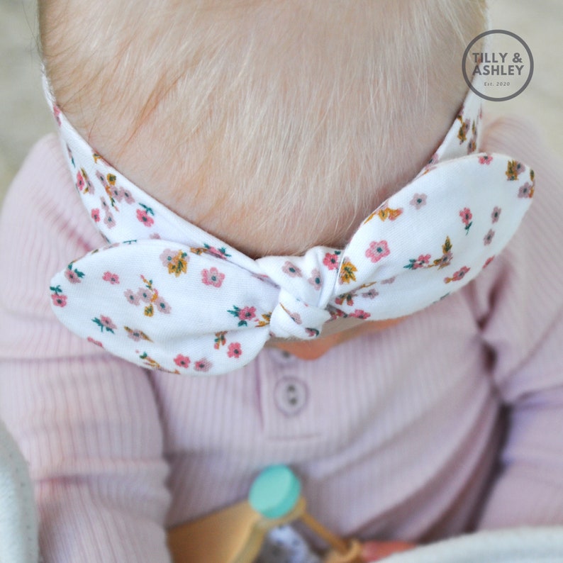 Puede incluir: Una diadema blanca con estampado floral y un lazo. La diadema est&aacute; atada alrededor de la cabeza de un beb&eacute; que lleva una camisa rosa.