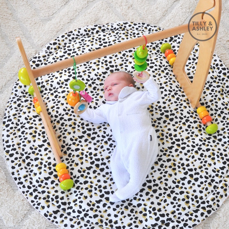 May include: A baby lies on a round play mat with a black and gold leopard print. A wooden baby gym with colorful hanging toys is above the baby. The text "Tilly & Ashley Est. 2020" is visible.