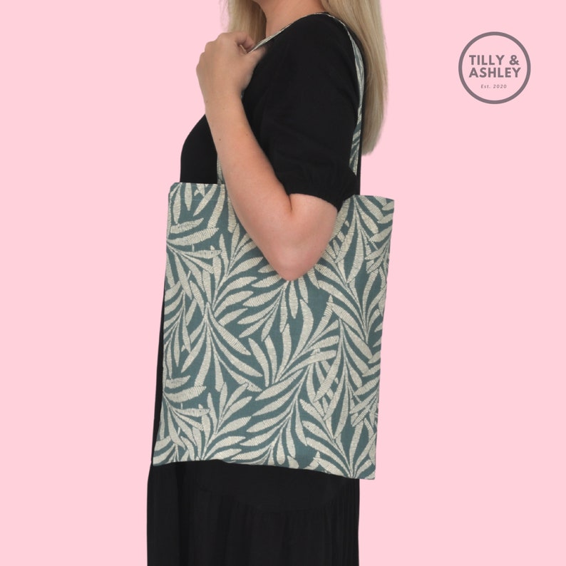 May include: A teal tote bag with a white leaf pattern. The bag has a long strap and is being held by a person. The bag has the text "TILLY & ASHLEY Est. 2020" on the top right corner.