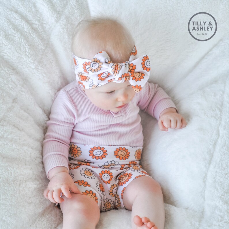 May include: A baby wearing a pink long-sleeved shirt and white shorts with a sunflower print. The baby is also wearing a white headband with a sunflower print and a bow.