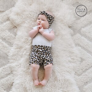 May include: A baby wearing a grey bodysuit with a leopard print waistband and a leopard print headband. The baby is lying on a white fluffy rug. The text "TILLY & ASHLEY Est. 2019" is in the top right corner of the image.