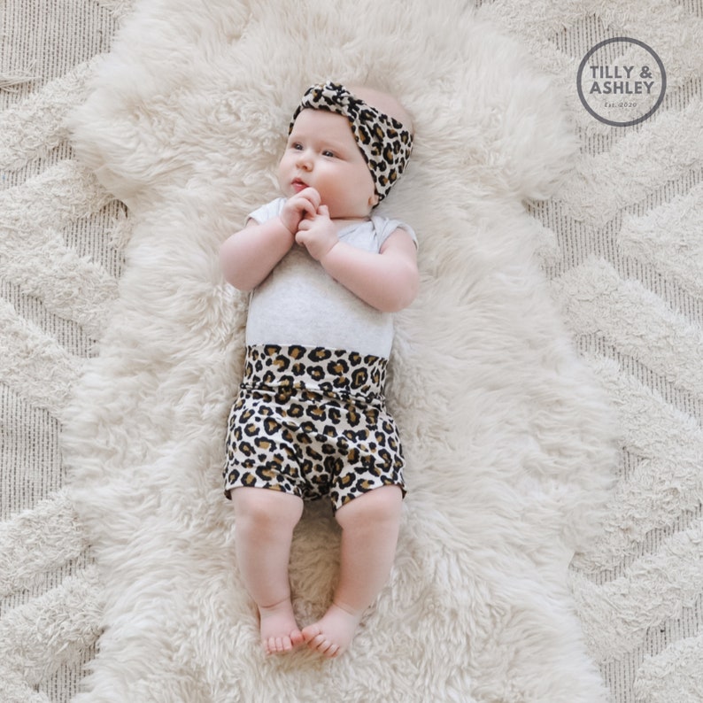 May include: A baby wearing a grey bodysuit with a leopard print waistband and a leopard print headband. The baby is lying on a white fluffy rug. The text "TILLY & ASHLEY Est. 2019" is in the top right corner of the image.