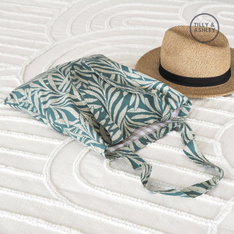 May include: A teal and white patterned tote bag with a white and black checkered lining. The bag is folded and lying on a white textured surface. A brown straw hat with a black band is lying on the surface behind the bag. The hat has a label that says "Tilly & Ashley".