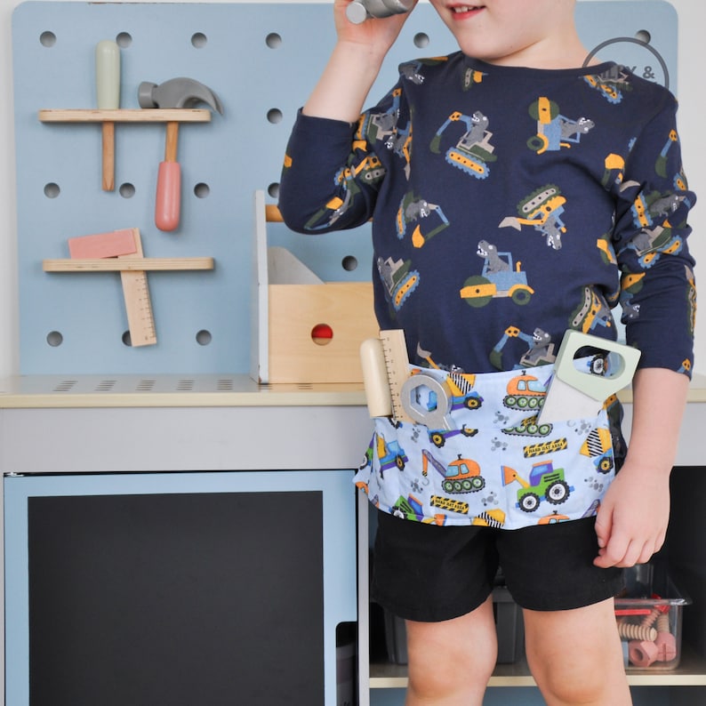 May include: A young child wearing a blue long-sleeved shirt with a construction vehicle print and a blue and white tool belt with a construction vehicle print. The child is holding a toy tool in one hand and is standing in front of a blue pegboard with various tools hanging on it.