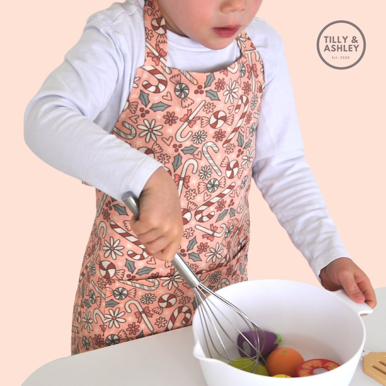 May include: A child wearing a pink and white candy cane and holly print apron is using a whisk to mix ingredients in a white bowl. The apron has a pocket on the front. The text "TILLY & ASHLEY Est. 2020" is on a white tag attached to the apron.