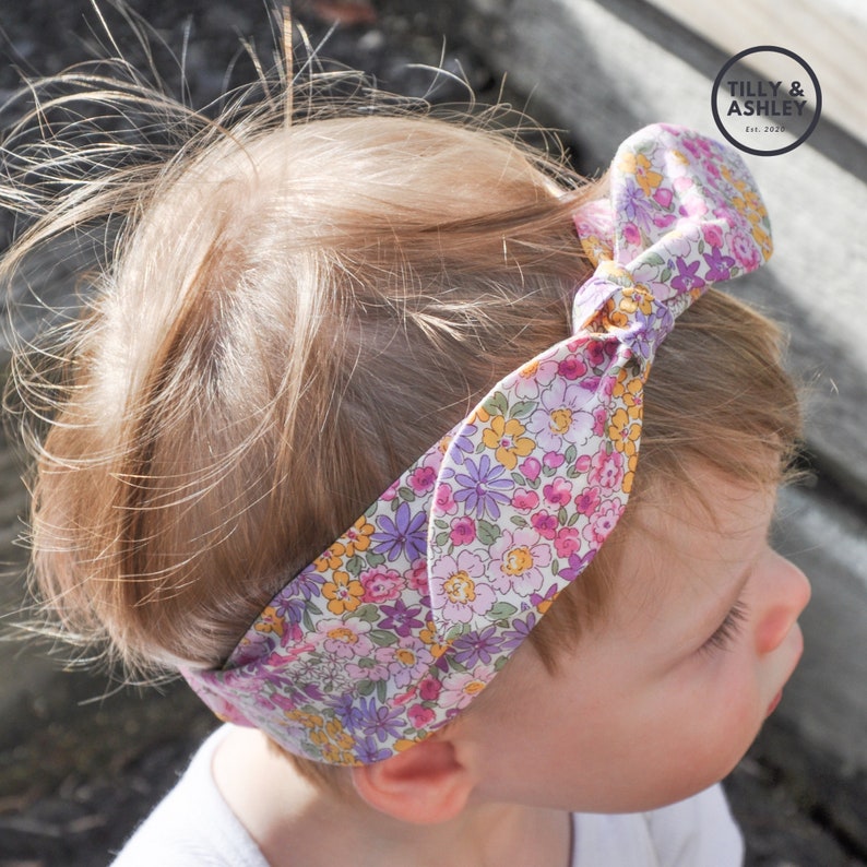 Puede incluir: Un ni&ntilde;o peque&ntilde;o con una diadema floral con un lazo en la parte superior. La diadema es blanca con flores rosas, moradas y amarillas. La diadema tiene el texto "TILLY & ASHLEY Est. 2019" en el lazo.