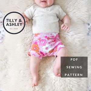 Puede incluir: Un patrón de costura PDF para pantalones cortos de bebé con estampado floral rosa y blanco. El patrón está disponible para descargar. Tilly & Ashley Est. 2020.