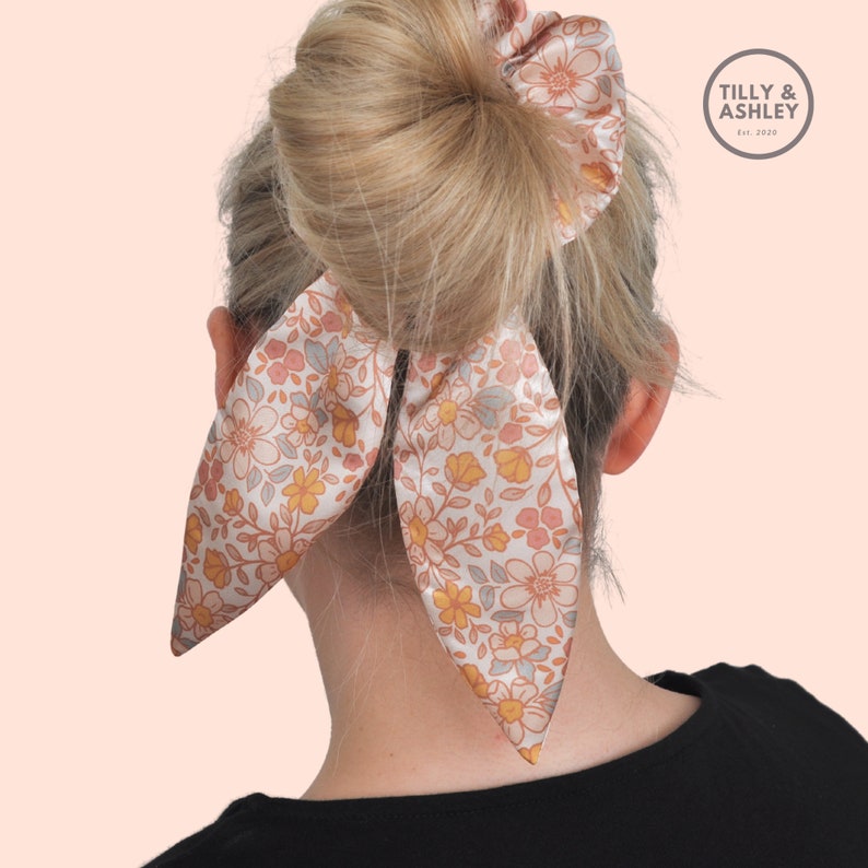 May include: A floral patterned hair scrunchie with a matching ribbon tied in a bow. The scrunchie is made of satin and has a light pink, yellow, and grey colour scheme. The ribbon is tied in a bow at the back of the head. The brand name "TILLY & ASHLEY Est. 2020" is visible in the top right corner of the image.