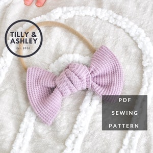 May include: A purple waffle knit fabric bow headband with a brown elastic headband. The headband is on a white fluffy surface. The text "TILLY & ASHLEY Est. 2020" is in a black circle above the headband. The text "PDF SEWING PATTERN" is in a black rectangle below the headband.