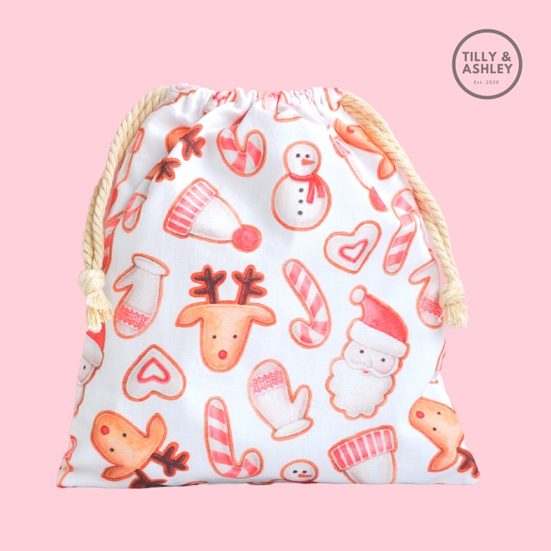 May include: White drawstring bag with a Christmas cookie pattern. The pattern includes gingerbread men, candy canes, mittens, reindeer, and hearts. The bag has a white drawstring and a white label with the text "TILLY & ASHLEY Est. 2019".