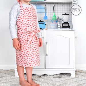 May include: A child wearing a pink and white apron with a strawberry print. The apron has a tie at the waist. The child is standing in front of a white toy kitchen.