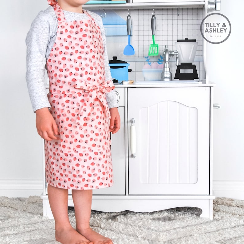 May include: A child wearing a pink and white apron with a strawberry print. The apron has a tie at the waist. The child is standing in front of a white toy kitchen.