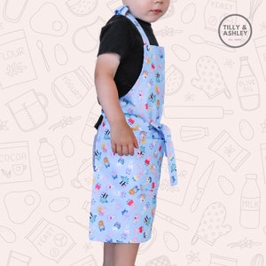May include: A child wearing a blue and white apron with a cartoon character print. The apron has a pocket and adjustable straps. The apron is made by Tilly & Ashley, a company established in 2020.