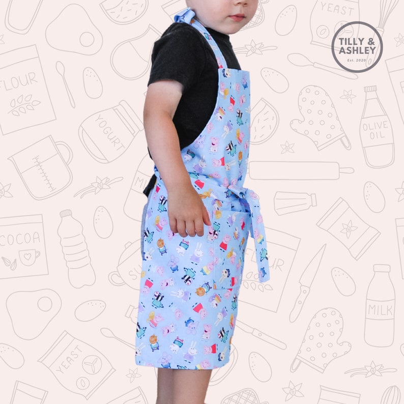 May include: A child wearing a blue and white apron with a cartoon character print. The apron has a pocket and adjustable straps. The apron is made by Tilly & Ashley, a company established in 2020.