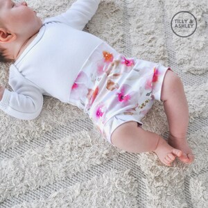 May include: A baby wearing a white long-sleeved onesie with a floral print bottom. The onesie has a white bodice and a pink, orange, and brown floral print bottom. The baby is lying on a white fluffy rug.