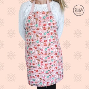 May include: Pink Christmas-themed apron with a pattern of gingerbread men, candy canes, and other holiday baking items. The apron has two pockets and adjustable straps. The text "TILLY & ASHLEY Est. 2020" is printed on a white tag.