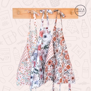 May include: Three floral print aprons with pink and white gingham trim hanging on a wooden rack. The aprons have a variety of floral patterns, including roses, sunflowers, and other wildflowers. The text "TILLY & ASHLEY est. 2020" is visible on the rack.