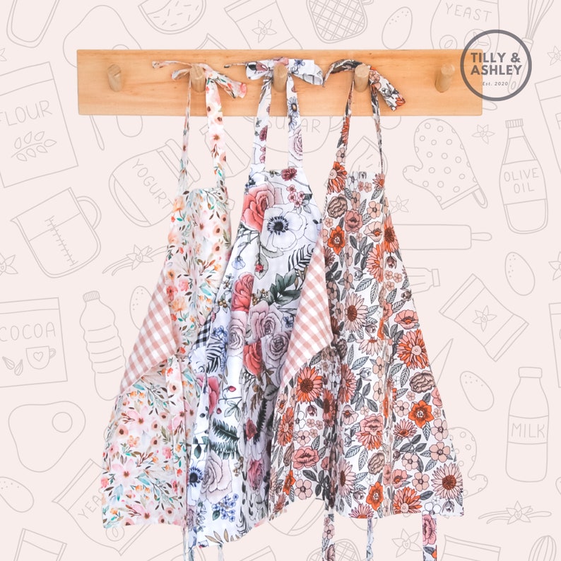 May include: Three floral print aprons with pink and white gingham trim hanging on a wooden rack. The aprons have a variety of floral patterns, including roses, sunflowers, and other wildflowers. The text "TILLY & ASHLEY est. 2020" is visible on the rack.