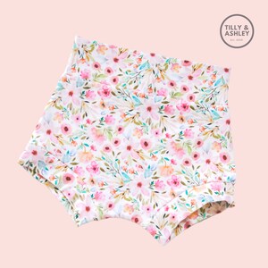 May include: White cotton baby bloomers with a pink and green floral print. The print features watercolor-style flowers and leaves.
