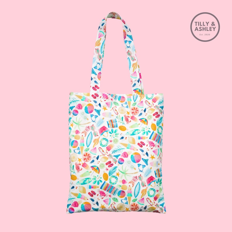 May include: White tote bag with a colorful summer-themed print featuring various beach items like flip flops, ice cream cones, watermelon slices, and palm trees. The bag has two handles and the text "TILLY & ASHLEY Est. 2019" is printed on a small circle in the top right corner.