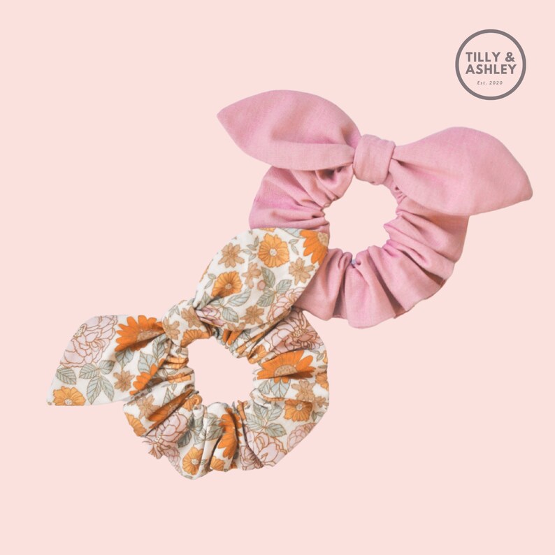 May include: Two fabric scrunchies, one with a floral print and one solid pink. The floral scrunchie has a bow and the pink scrunchie has a bow.